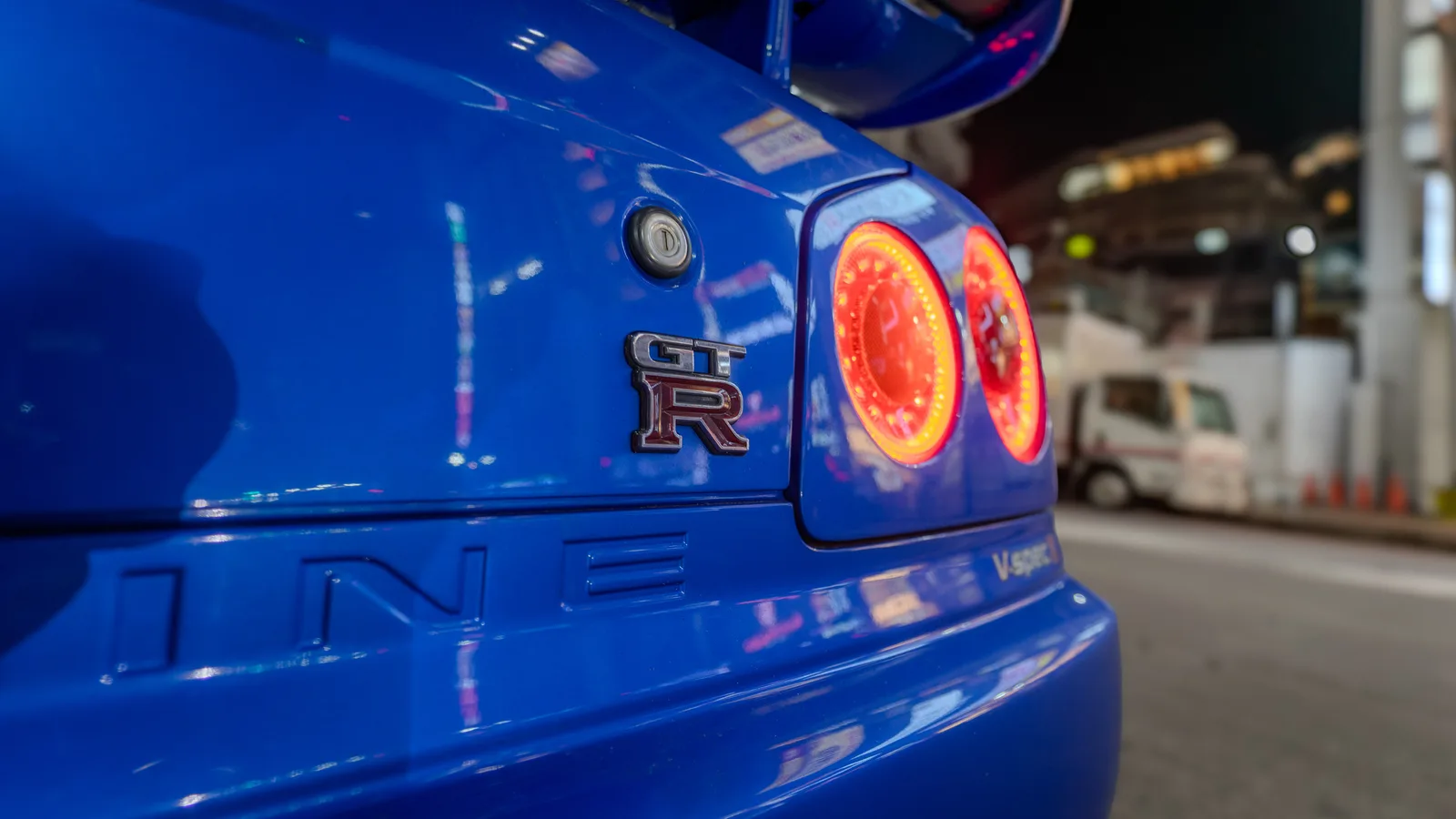 โลโก้ GT-R และไฟท้ายกลมของ R34 V-spec II สีน้ำเงิน ถ่ายกลางคืนในเมือง