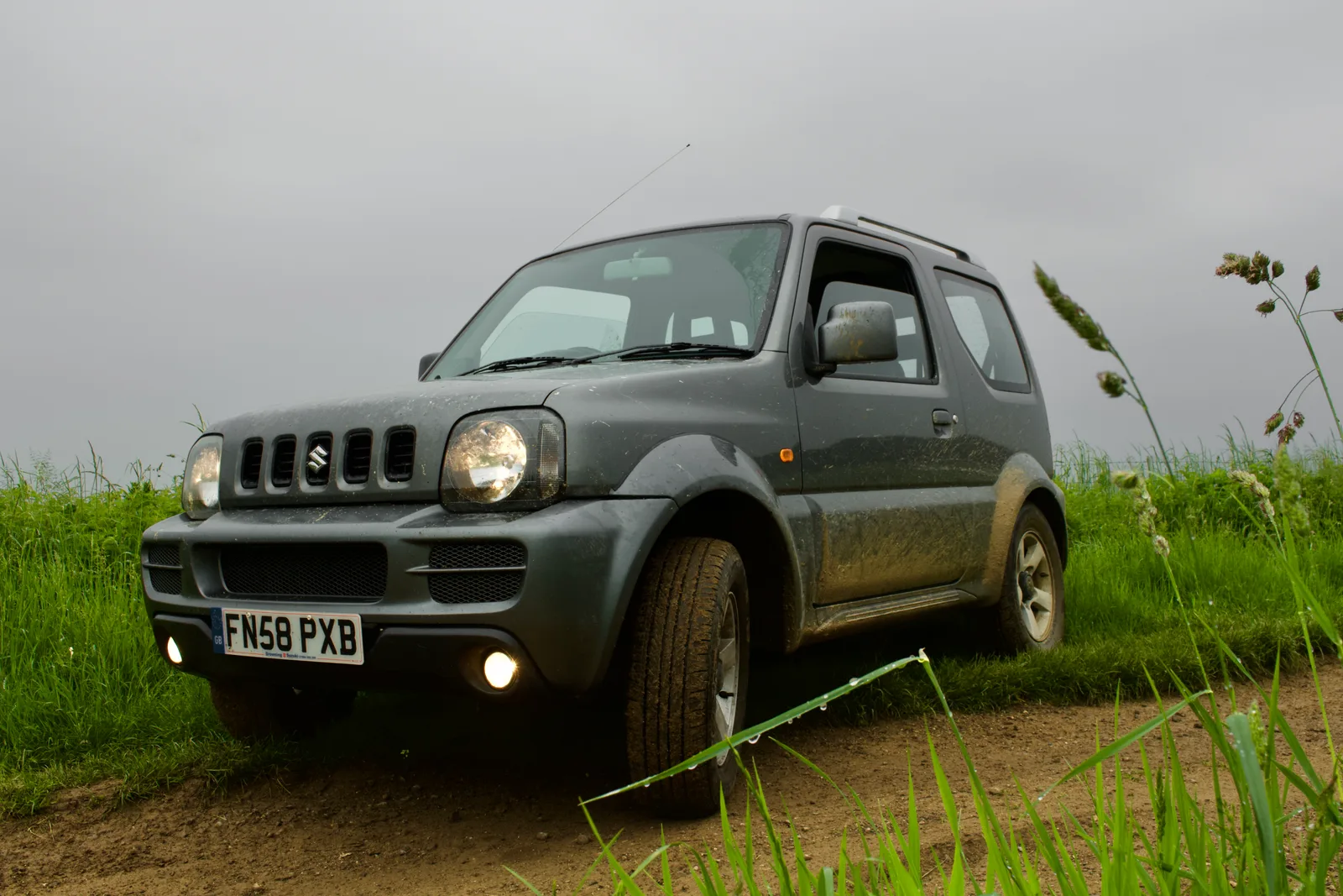 Jimny สีเทา มุมหน้าเตี้ย บนทางดินกลางทุ่งวันฟ้าครึ้ม