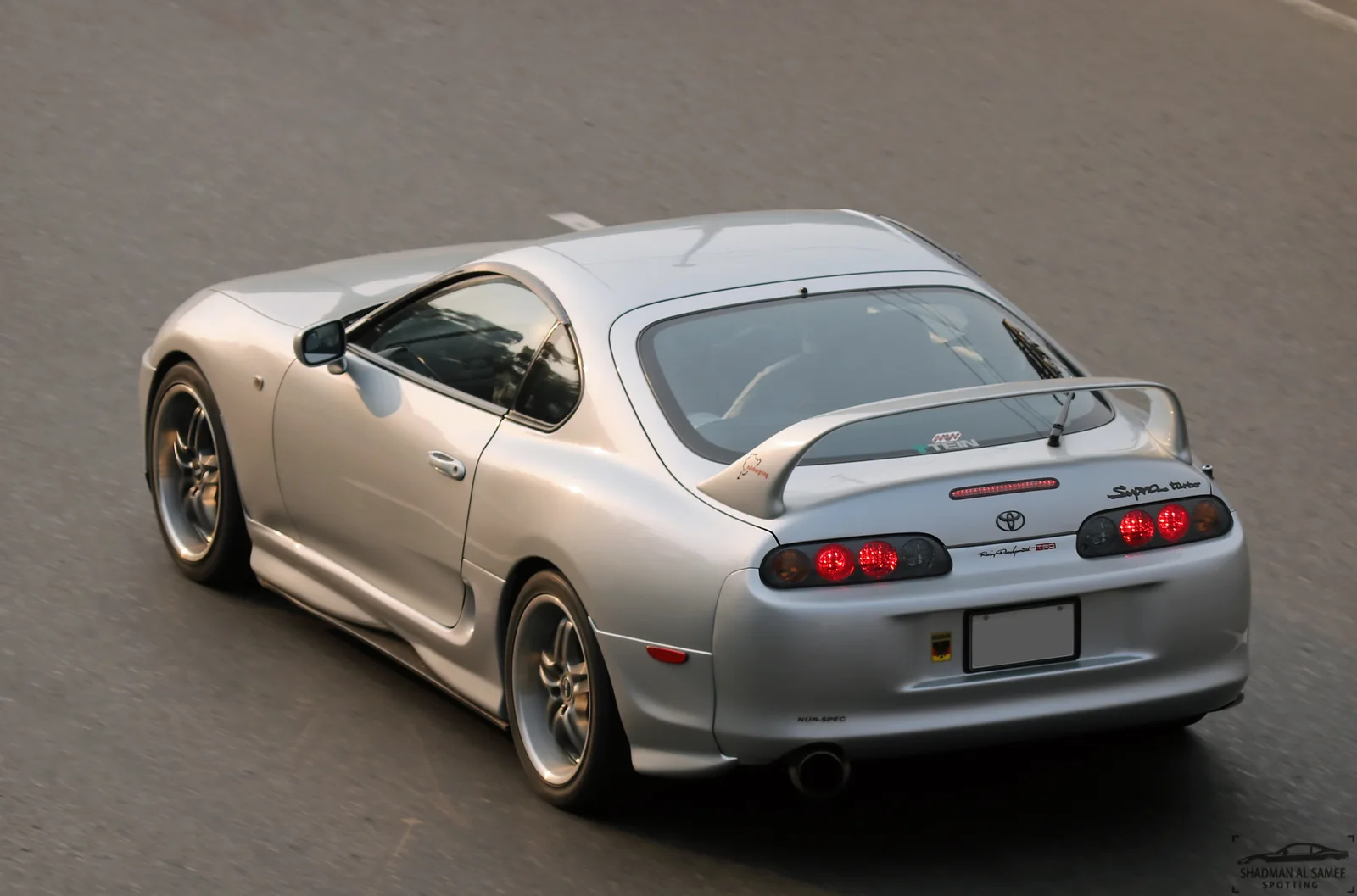 Toyota Supra JZA80 สีเงิน มุมท้ายสามส่วน กำลังวิ่งบนถนน ติดสปอยเลอร์ใหญ่ชัดเจน