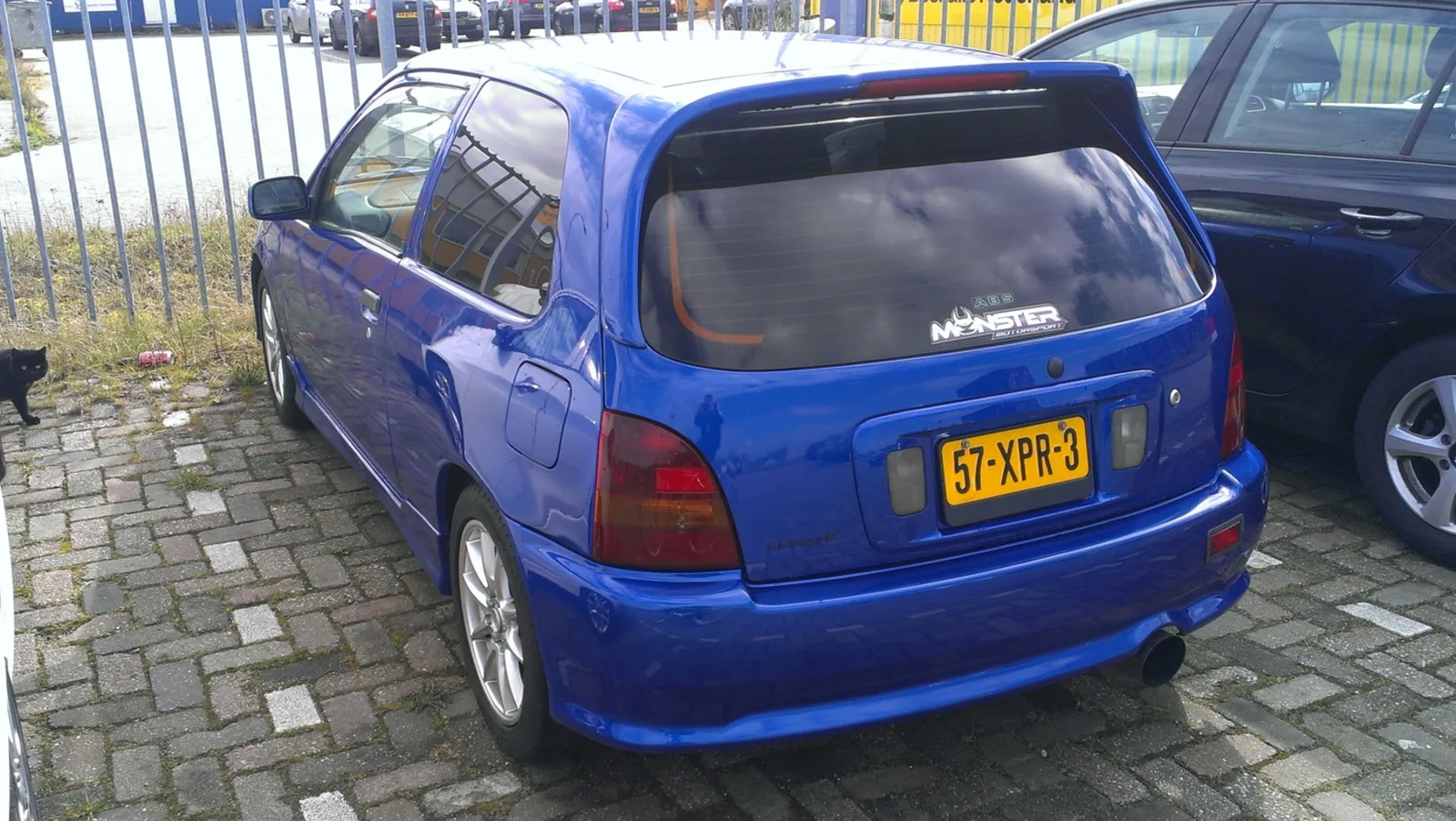 Glanza V สีน้ำเงิน มุมท้าย ติดสติกเกอร์ Monster Motorsport จอดริมรั้วเนเธอร์แลนด์