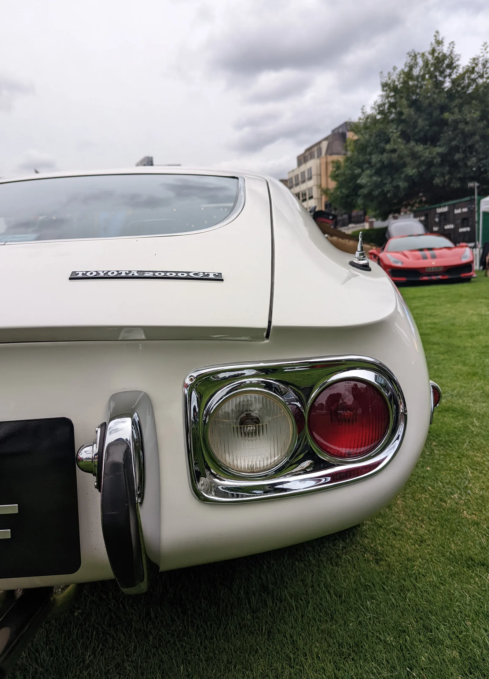 ดีเทลไฟท้ายโครเมียมและแบดจ์ Toyota 2000GT กลางแจ้ง หลังรถมีเฟอร์รารีสีแดงเบลอ