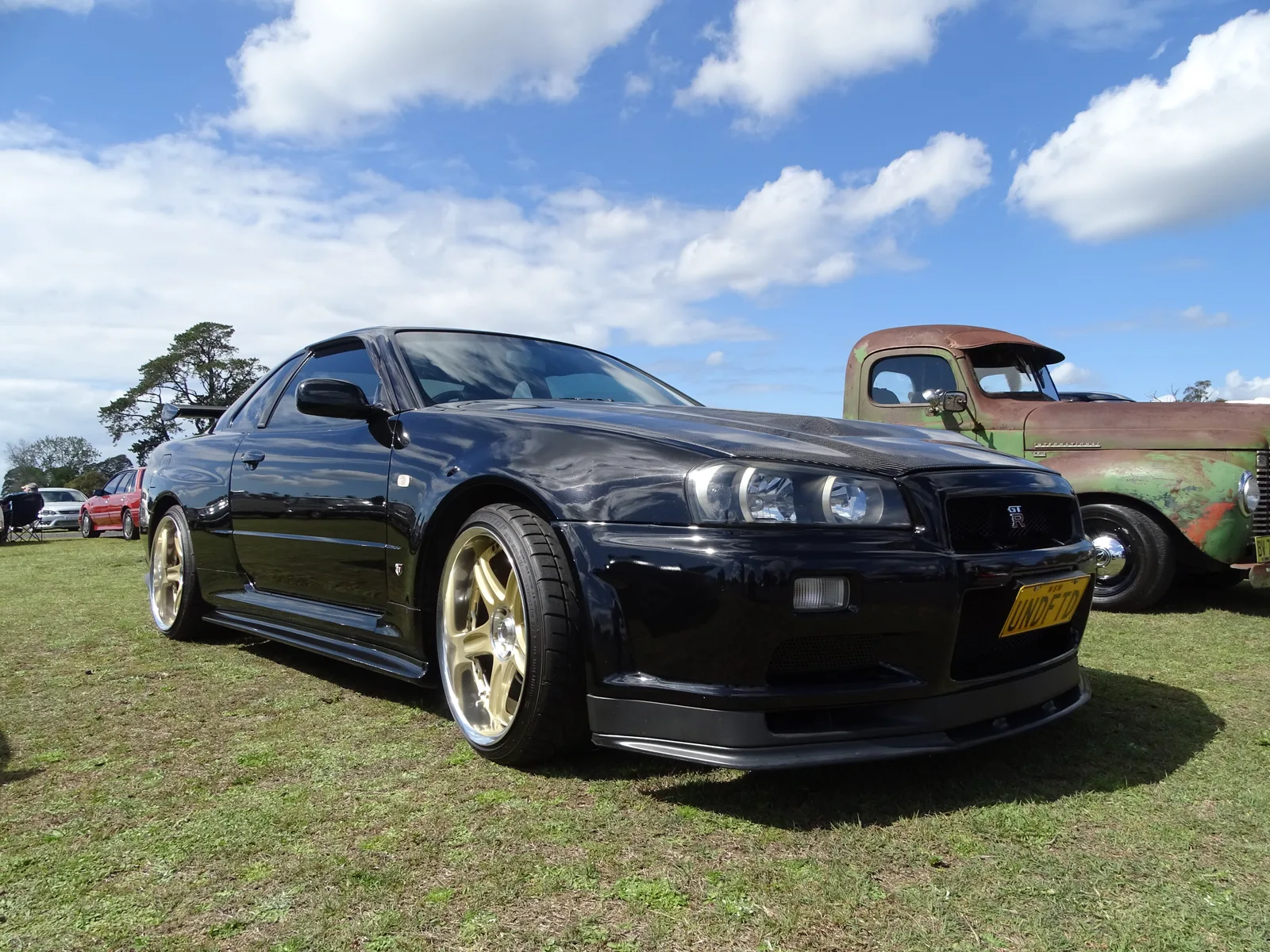 Skyline GT-R R34 V-spec II สีดำ ล้อทอง มุมหน้า 3/4 ใต้ฟ้าเปิด