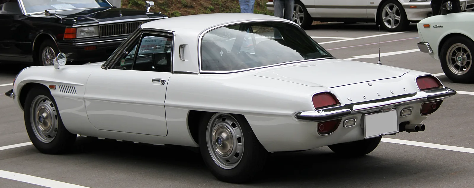 Mazda Cosmo Sport สีขาว มุมข้างท้ายเฉียง จอดกลางแจ้งท่ามกลางรถคลาสสิกคันอื่น