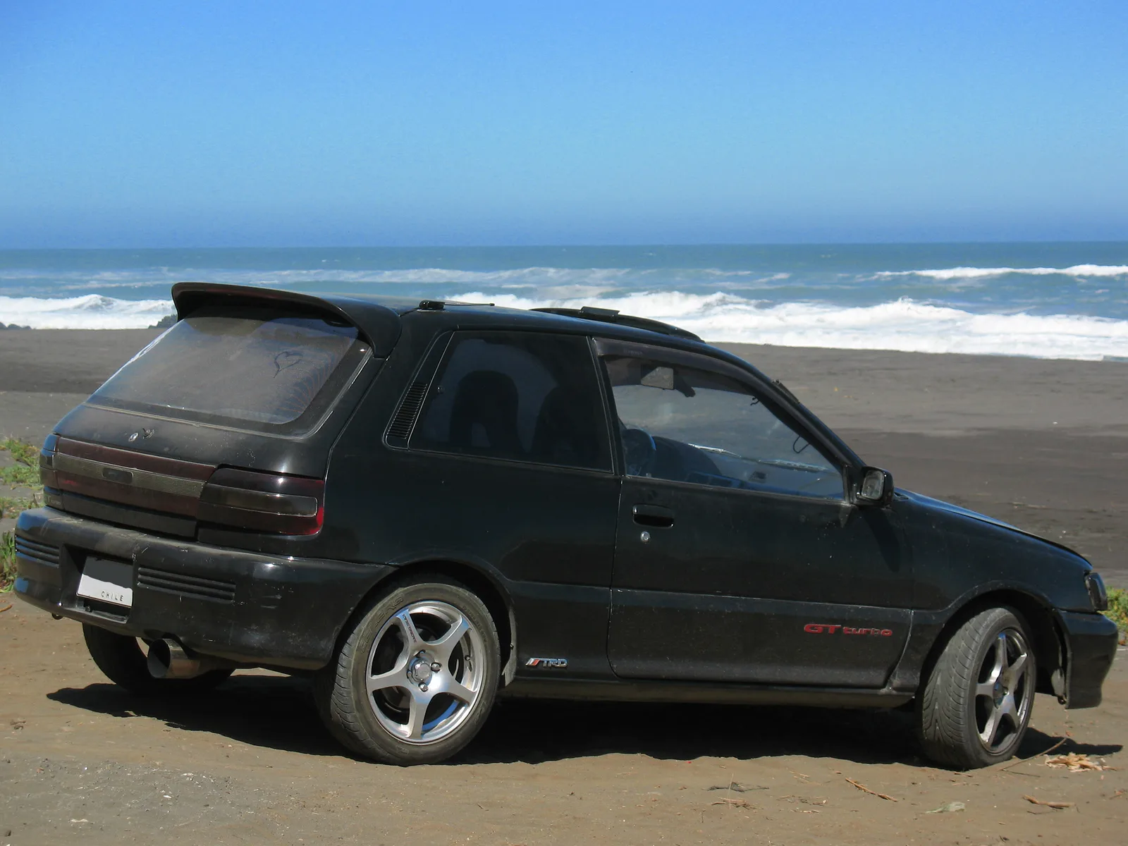 Starlet GT turbo EP82 สีดำ มุมท้ายเฉียง จอดริมหาด เห็นสปอยเลอร์และล้อแต่ง