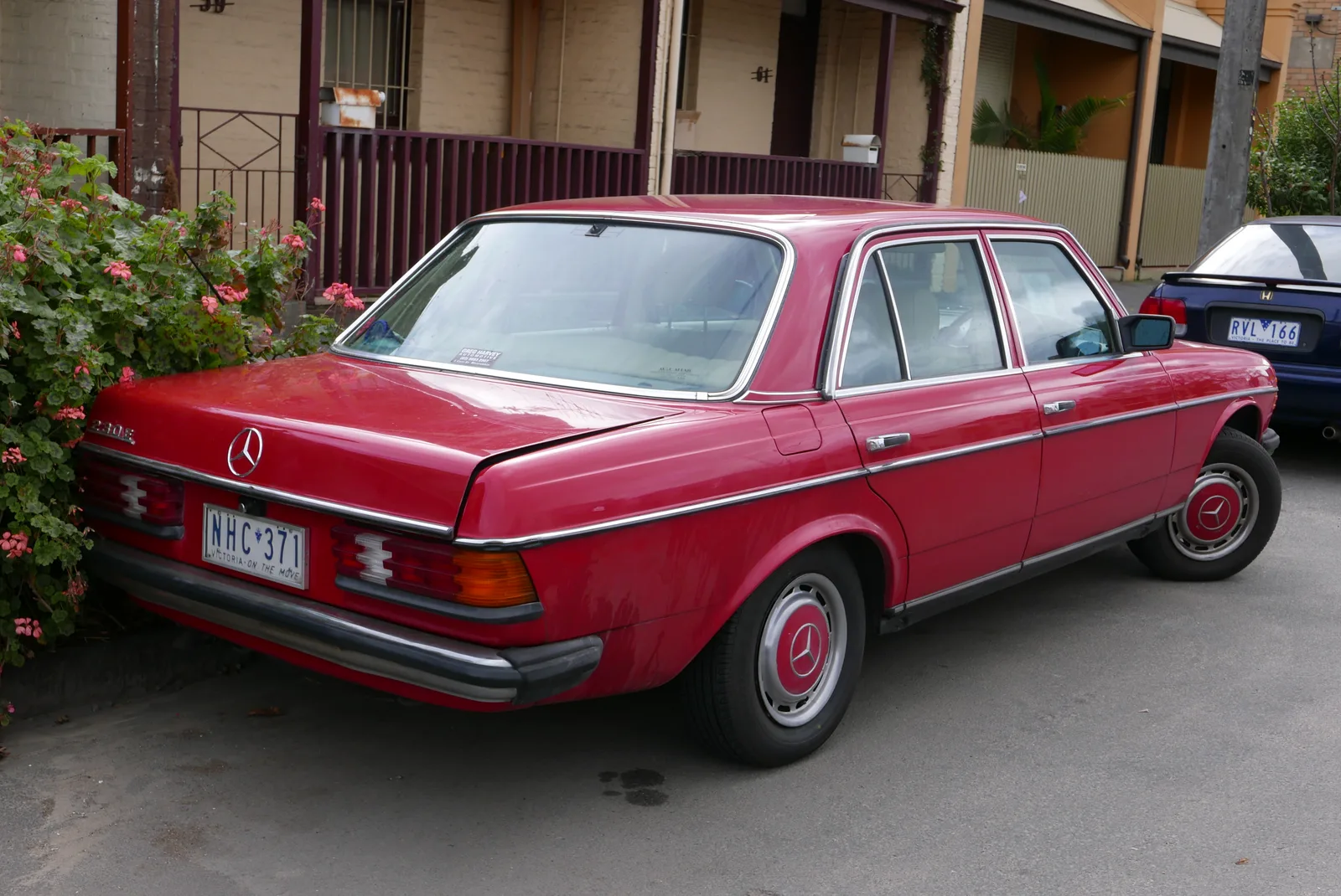 W123 สีแดงสด มุมท้ายเฉียงจอดริมถนนย่านบ้านพัก สีสันสดใสตัดกับพุ่มไม้