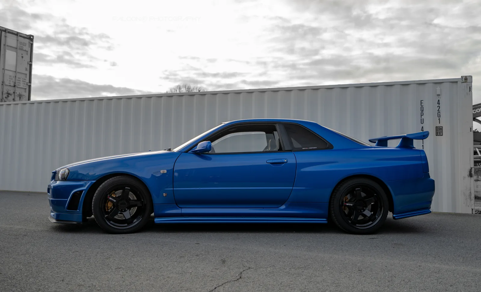 R34 GT-R สี Bayside Blue มุมข้างเต็มคัน หน้าตู้คอนเทนเนอร์