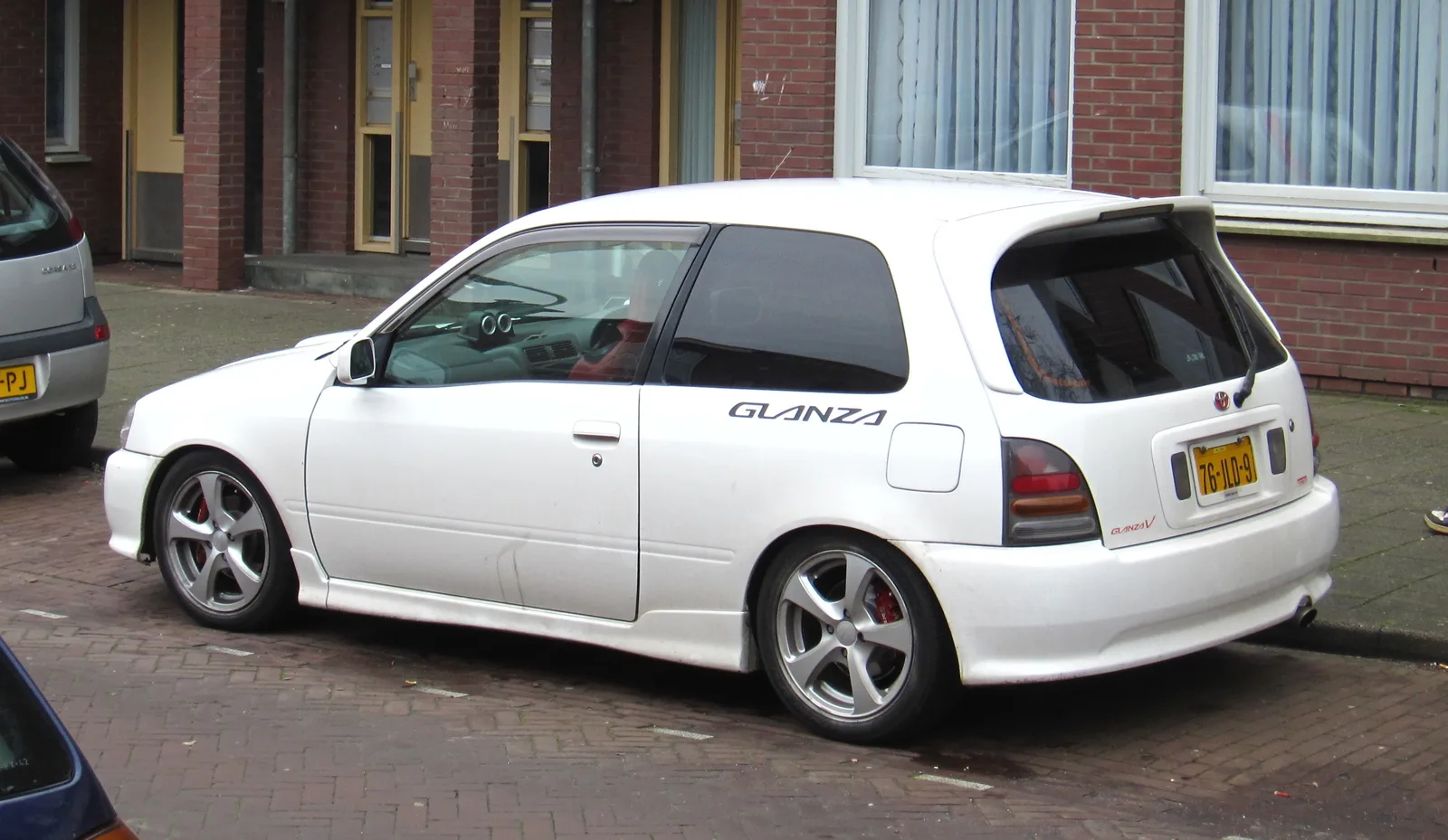 Glanza V สีขาว มุมข้างเต็มคัน แม็กแต่งและคาลิเปอร์แดง จอดริมตึกอิฐยุโรป