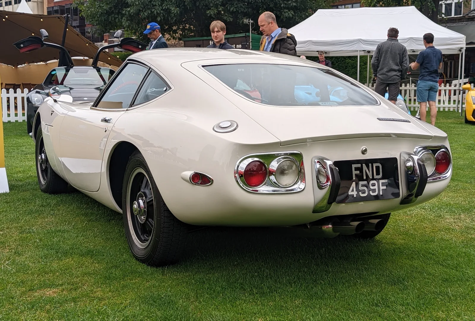 Toyota 2000GT สีครีม มุมท้ายเฉียง ในงาน Concours กลางสนามหญ้า มีผู้ชมรายล้อม
