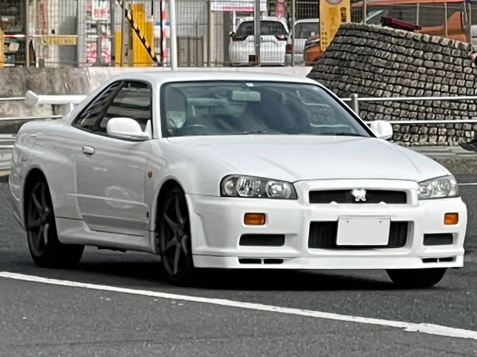 R34 GT-R สีขาววิ่งบนถนนญี่ปุ่น มุมหน้าสามส่วน เห็นกันชนหน้าและไฟหน้าทรงกลม