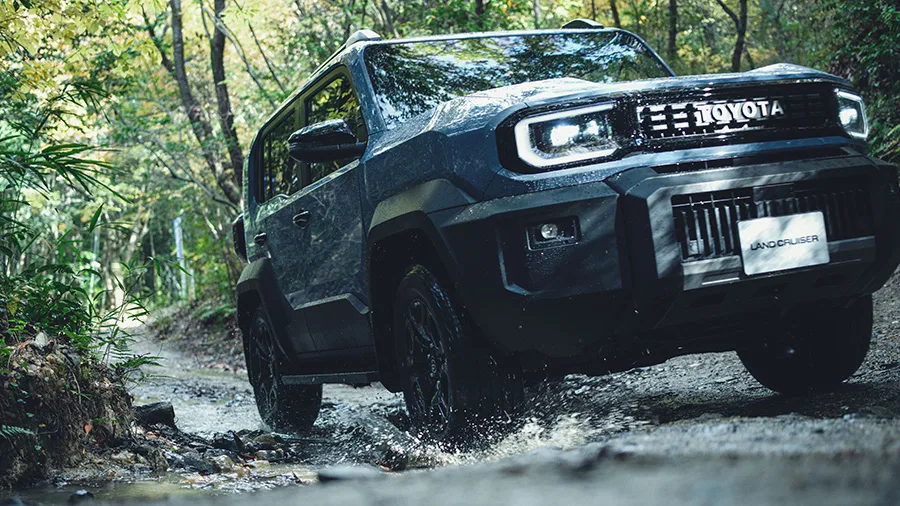 Land Cruiser FJ สีเข้ม ลุยน้ำบนทางป่า มุมหน้าสามส่วนต่ำ เห็นไฟหน้า LED และกระจังโลโก้ TOYO