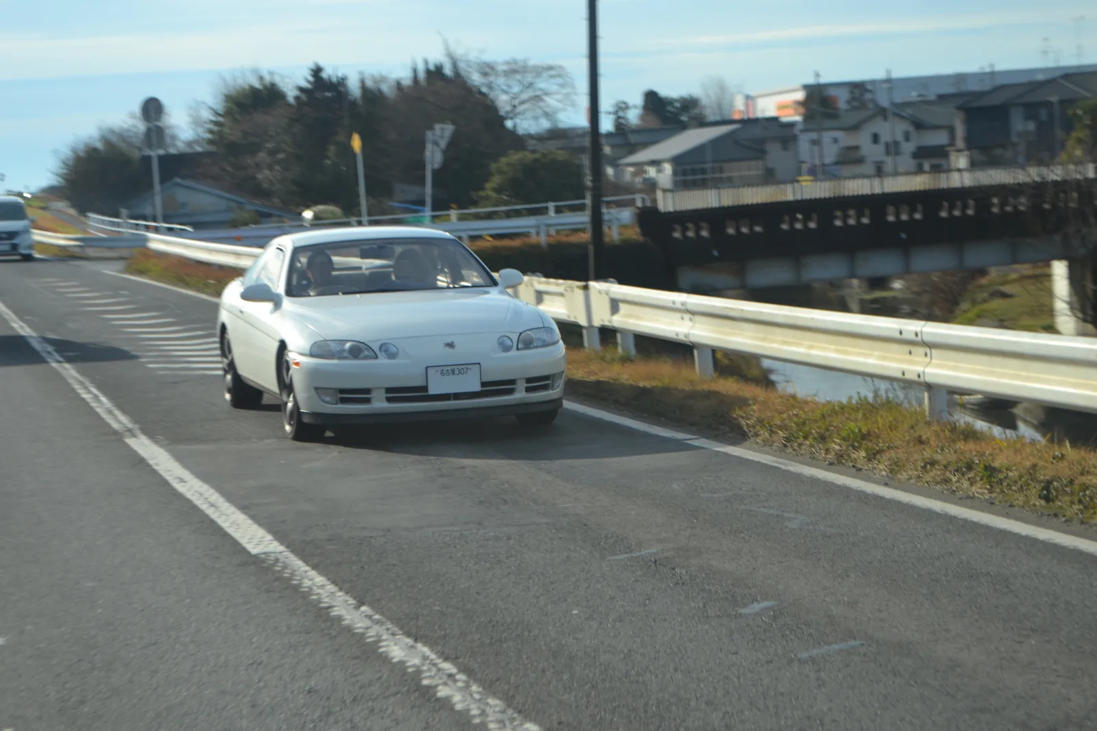 Soarer Z30 สีขาวมุก กำลังวิ่งบนถนนริมคลองในญี่ปุ่น มองเห็นหน้าตาไฟหน้ารุ่นนี้ชัด