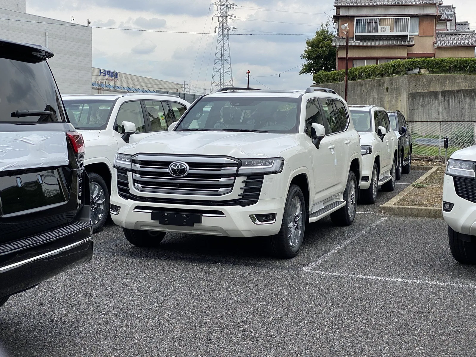 Land Cruiser 300 สีขาว หน้าตรงในลานจอดตัวแทนจำหน่ายที่ญี่ปุ่น ยังติดเทปกันรอยบนฝากระโปรง