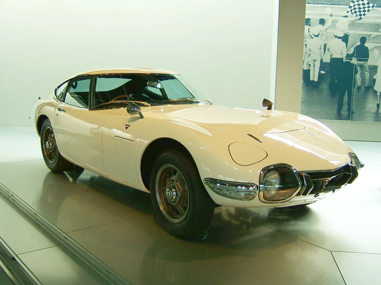 Toyota 2000GT สีขาวครีม หน้าสามส่วน จอดโชว์ในพิพิธภัณฑ์ พร้อมภาพธงหมากรุกด้านหลัง